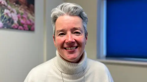 Jane St Pier - Headshot of a lady with short grey hair smiling in a white turtleneck jumper