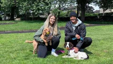 A woman and man kneeling on grass in a park with two spaniel dogs