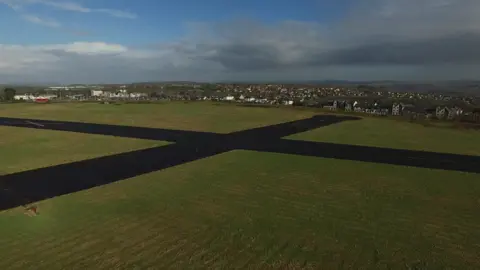 An aerial image of an airport runway in the middle of green grass with buildings in the background