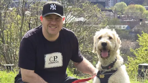Ray Clements, who wears a black baseball cap and t-shirt, smiles as he crouches down next to his dog. They are on a slope with trees and urban buildings behind.