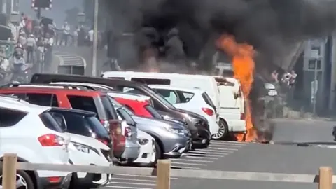 Flames are erupting from a white van and plumes of black smoke are going up into the sky. The van is parked in a row of other cars and people can be seen in the background leaving the scene.
