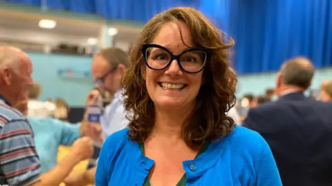 A head-and-shoulders shot of Sarah. She has shoulder length brown hair, is wearing large glasses and a light blue cardigan.