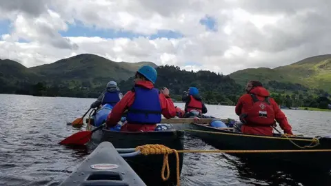 Four people, viewed from behind, canoeing on a lake. Green hills can be seen beyond the water.