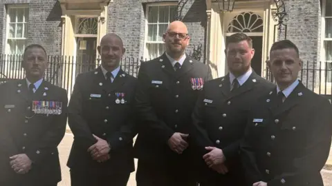 XPR Communications Ltd A row of five male police officers in dark dress uniforms standing in a line outside No 10 Downing Street. They all have their hands folded in front of them. 