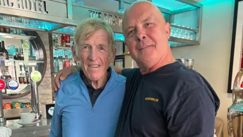 Don Taylor An older man with short blonde and grey hair and a light blue top is smiling while standing next to another older man with a bald head and a dark blue T-shirt. They are standing in a bar area.