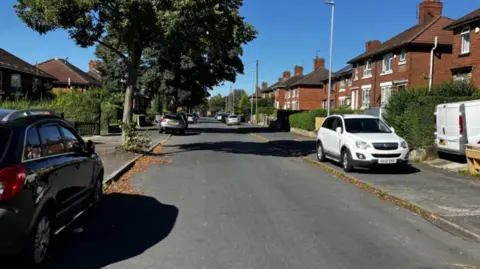 A residential street with houses on either side, and cars parked on the pavement