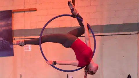 A circus school pupil hanging upside down on an aerial hoop inside a large hall.