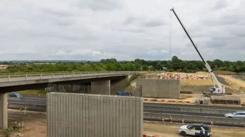 National Highways A grey concrete bridge over a dual carriageway. On either side of the dual carriageway construction is under way. The far side is busy, with a white crane and piles of road building materials. The near side is coned off but no work is being undertaken.