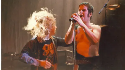 Des Rumney Des Rumney with long blonde hair, goes to hug Ozzy Osbourne on stage. Ozzy is shirtless as he is singing into a microphone.