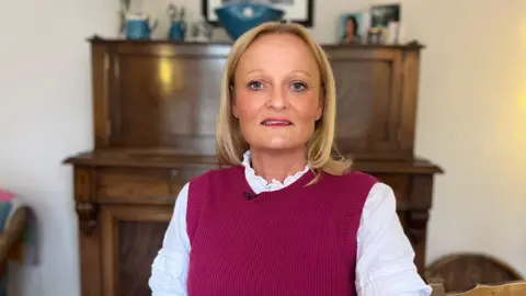 Maria Mulgrew looking to camera. She is sitting in front of a piano and is wearing a dark pink tank top and a white blouse with a frilled collar