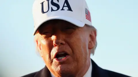 Trump pictured on 13 March. It is a close-up shot of his face in front of a blue sky. He wears a large white baseball cap with USA in large black letters on the front and a US flag on the side.