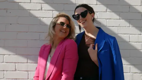 Two stylish women, one in a pink coat and another in a blue one. They both wear sunglasses and lean into each other.