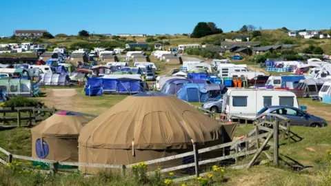 A British campsite, with caravans, cars, tents and a yurt scattered around a field.