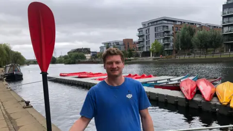 John Devine/BBC Byron Cripps standing with an oar near some boats