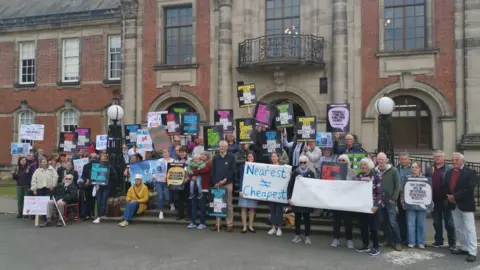 Local Democracy Reporting Service A group of people standing outside a large building, holding colourful banners. Some banners say 'bring catchment back' and 'save our schools'.