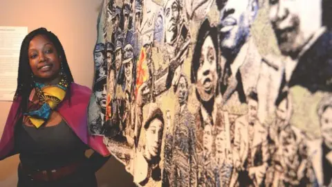 Paula Ogun Hector Ms Hector stands to the left of the tapestry, looking on at it. She has a bright pink jacket on, a blue and yellow scarf and wears braids. The tapestry is blue and yellow in colour and features different faces.