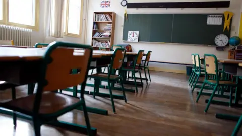 A generic shot of a classroom with chairs lined up against individual desks. At the back of the room, there is a long blackboard on the wall next to a bookshelf. 