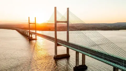 The Prince of Wales Bridge at sunrise. The river can be seen under the bridge and the orange sun is rising over the horizon behind it