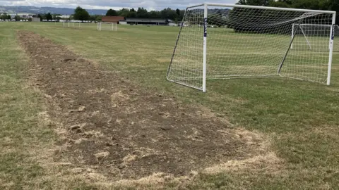 Football pitches with goalposts. A trench, which has been filled in, runs along the left of the image. There is a building and a stand in the background.