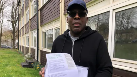 A close up of Joseph, wearing a blue baseball cap and sunglasses, standing outside the block of flats in Luton that is in need of fire safety work. His quarterly service and sinking fund bill is set to increase by more than 300% from April