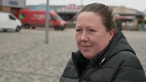 A woman with brown hair, tied back, stands in a car park in front of seaside shops with two vans, one white, one red, parked in the background. She is wearing a black coat.