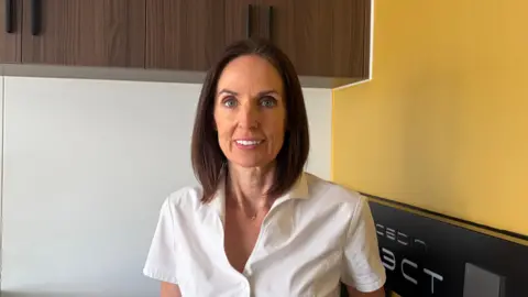 Ruth Alexander has shoulder-length dark hair smiles at the camera. She is wearing a white top, and is standing next to a yellow wall.