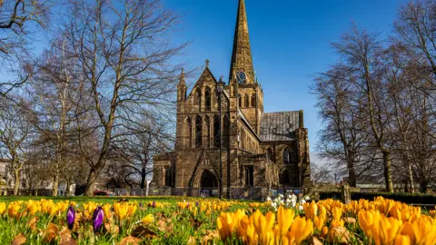 BBC Weather Watchers/DarloMur A large gothic church surrounded by trees without leaves. In the foreground there is a patch of grass in the foreground with yellow, purple and white flowers. The sky is bright blue with no clouds.