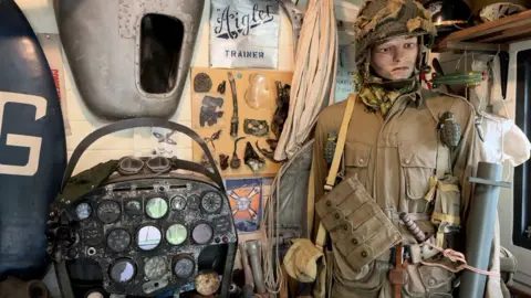 A mannequin dressed in World War Two army uniform complete with grenade stands in a shed. Next to it are various other World War Two memorabilia including a dashboard from a plane, weaponry and posters.