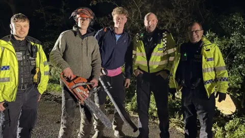 Five people, stood in a line, facing the camera. There are three police officers all in uniform, and high-vis jackets, and two young men wearing dark clothing. One is holding a chainsaw and the other a leaf blower. Foliage is behind them. 