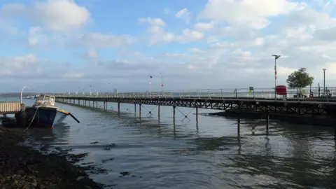 Picture shows a long pier stretching into distance with water in the foreground and a wrecked boat to the left.