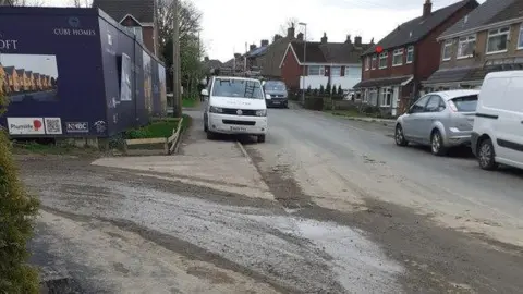 Wendy Cash The entrance to the housing development, with what looks like mud and water going from the edge of the road into the entrance. There are boards at the site entrance saying Cube Homes and including some computer generated images of some of the properties. There are houses over the road, with some cars parked and a van parked half on the pavement and half on the road.