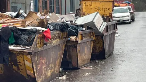 Geoff Howarth Three yellow skips overflowing with waste including furniture