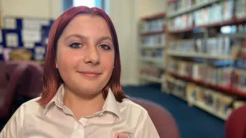 A girl called Millie is smiling at the camera. She has red hair that is shoulder length. She is wearing a white shirt. 