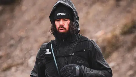 Mike Humphreys running in the rain. He is wearing a black hat, a black coat with his hood up, a black running vest and black gloves. 