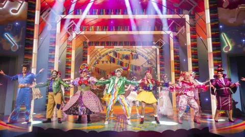 Pamela Raith A photo showing a stage with a number of performers taking part in a pantomime. They are all wearing colourful outfits