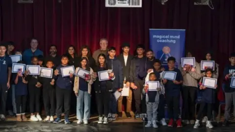 Nurture Media Young participants and mentors and Jin stand on a stage holding certificates. There is a large maroon curtain behind them. There are more than 20 people in the photo and a blue Magical Mind Coaching banner behind him.
