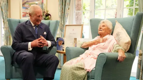 PA Media King Charles wearing a suit while sat in a green chair in a nursing home room. He is sat looking and smiling at an elderly woman, sat on his right, also in a green chair