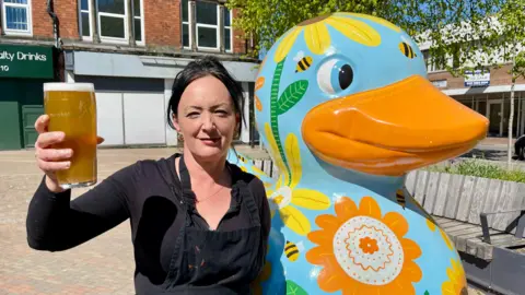 A woman with black hair, a black shirt and a black apron is holding a pint of lager aloft. She is stood beside a giant painted duck win a town square. The duck is blue, with an orange beak with flowers and bees painted on it. There are shops, benches, a tree and flower beds in the background.
