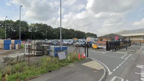 A Google maps street view of Swindon Household Waste Recycling Centre on the Cheney Manor Industrial Estate. There is a car park with lots of cars in it and buildings in the distance. Scrap metal and wood can be seen opposite the car park. 