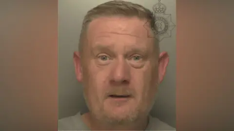 A custody image of Lawrence Worville who is wearing a grey sweatshirt as he stands against a grey background. He has light blue eyes, a beard and short light brown hair.