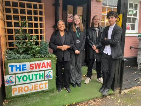 BBC/Amy Holmes A picture of a middle aged woman wearing black, a teenage girl and two teenage boys, who are also all wearing black, outside The Swan Youth Project in Berkhamsted.