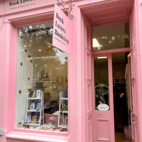 Caden Armstrong Book Lovers Bookshop from the outside. It has doors to the right and a window to the left and is painted in light pink. A sign also in pink with Book Lovers Bookshop A Romance Bookshop written in black hangs from a pink metal bracket. Two A-shaped small bookshelves with books are on display in the window and there are leaves attached to the inside. People can be seen inside the door reading books.