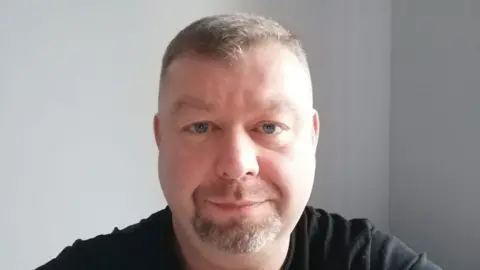 Tomasz Zareba A headshot of a man with short light hair and a goatee beard, He wears a black tshirt and stares into the camera.