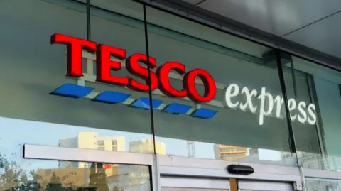 Getty Images A Tesco Express sign, from a generic store, on glass above the entrance doors. "Tesco" is in red with blue dashes below it, and "express" is in white. 