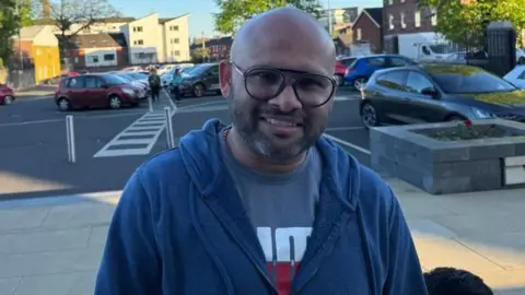 Blesson is msiling astatine  the camera. He has achromatic  framed glasses and is wearing a navy zip up   hoodie and grey t-shirt. 