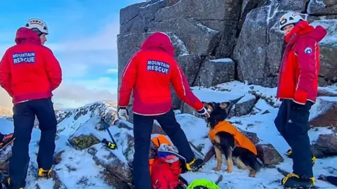 NNPMRT Three people with mountain rescue coats on a snowy outcrop with the dog Bramble wearing an orange coat seated at their feet 