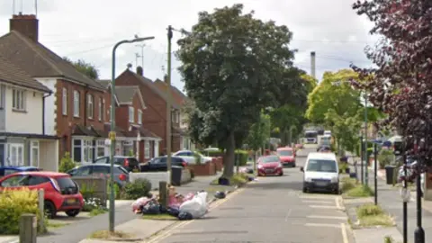 The street scene in Colemans Avenue, Southend-on-Sea. It is lined with houses and has cars parked outside them on driveways and along the road. There are trees also lining the road.