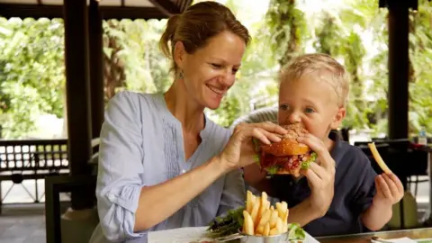 A woman with light brown hair feeds a burger to a boy