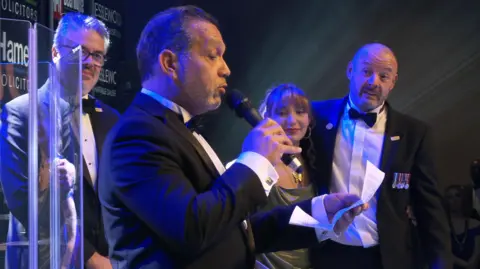The image shows a formal event on stage with bright spotlights. Several people in tuxedos stand near a podium, one holding a microphone and reading from a sheet of paper. Another man with medals on a jacket stands beside a young woman in a metallic gown. Sponsor logos are visible on a dark backdrop, and blue and gold lighting creates a dramatic atmosphere.
