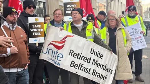 A group of protesters stand together in the street. Some of them are wearing hats that say defend the winter fuel payment and some others are wearing high-viz jackets. A few are holding signs.